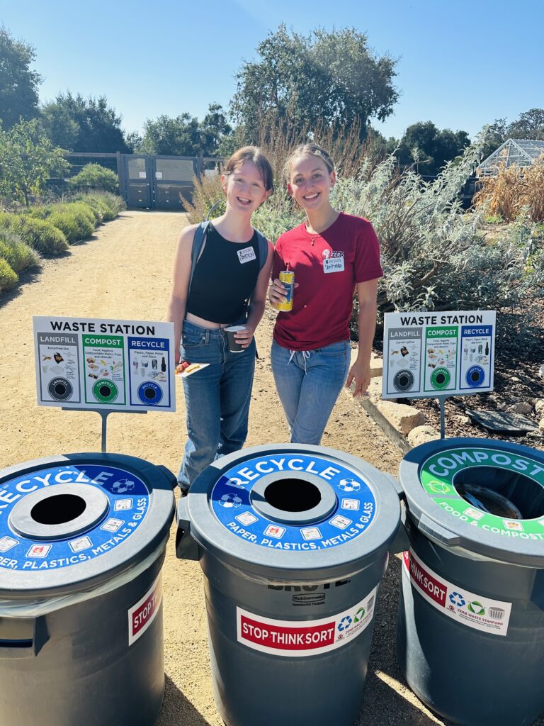 Two students standing behind recycling and composting bins