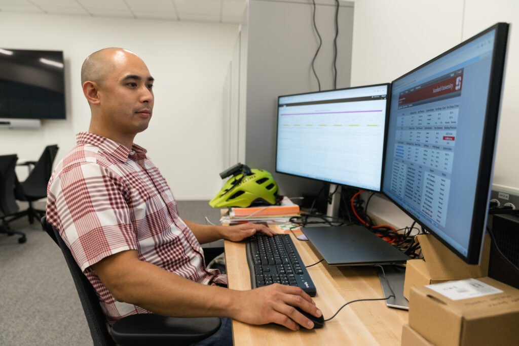 Man sitting at desk with two computer screen in front of him with one hand on the keyboard and the other on the mouse