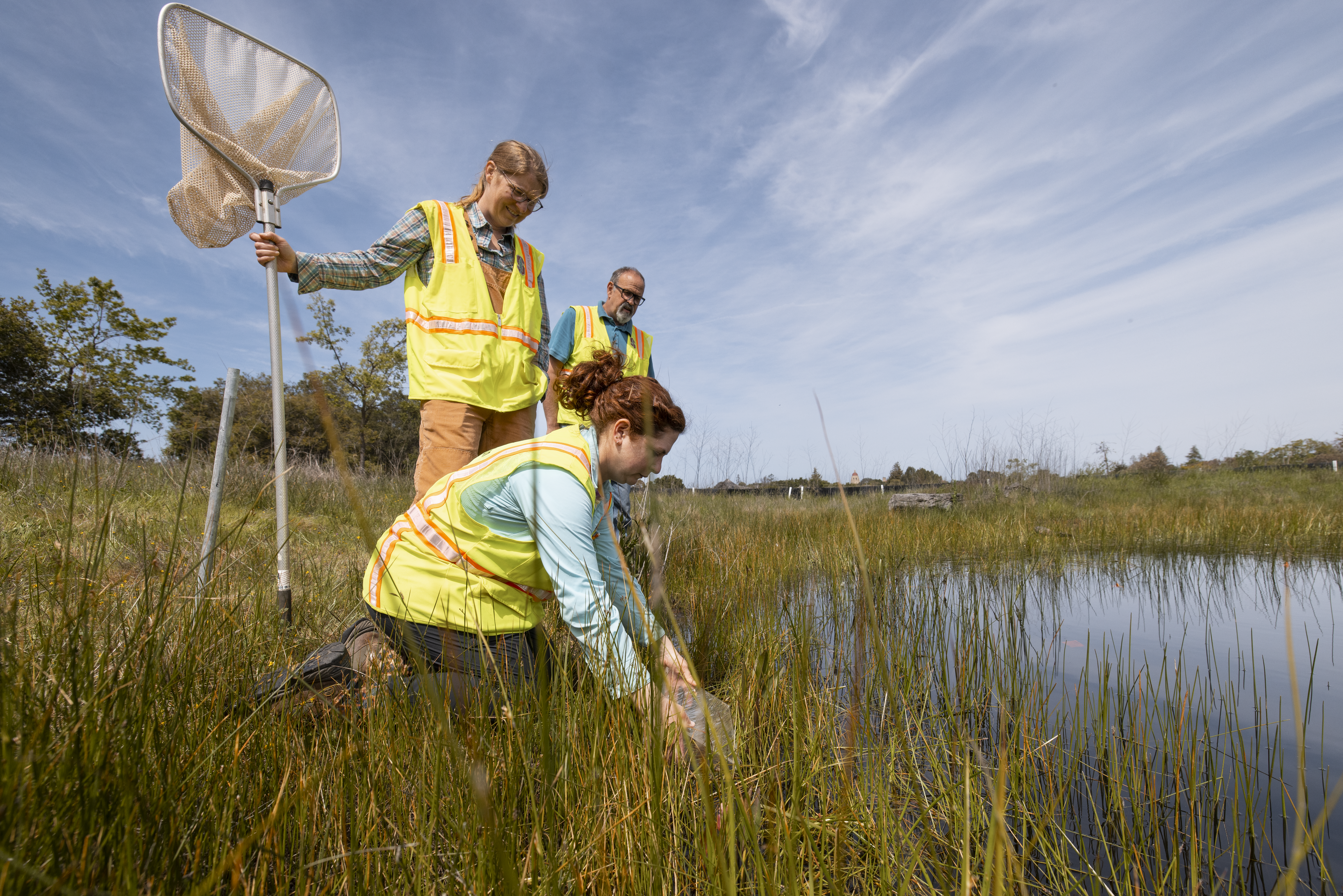 Person kneeling down by a body of water and two people standing up behind, one holding a net