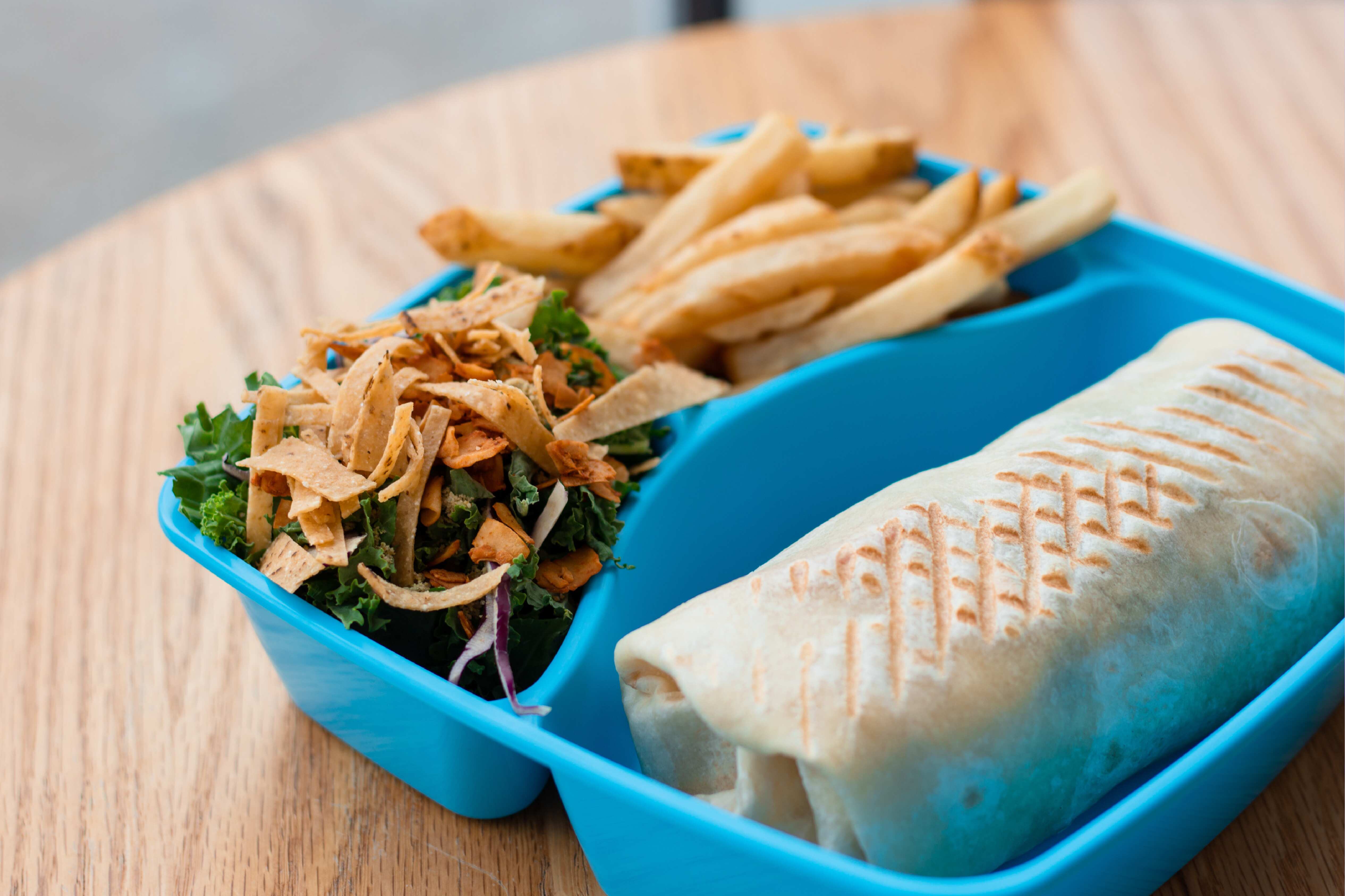 A blue to-go container on a table with a burrito, fries, and a salad.