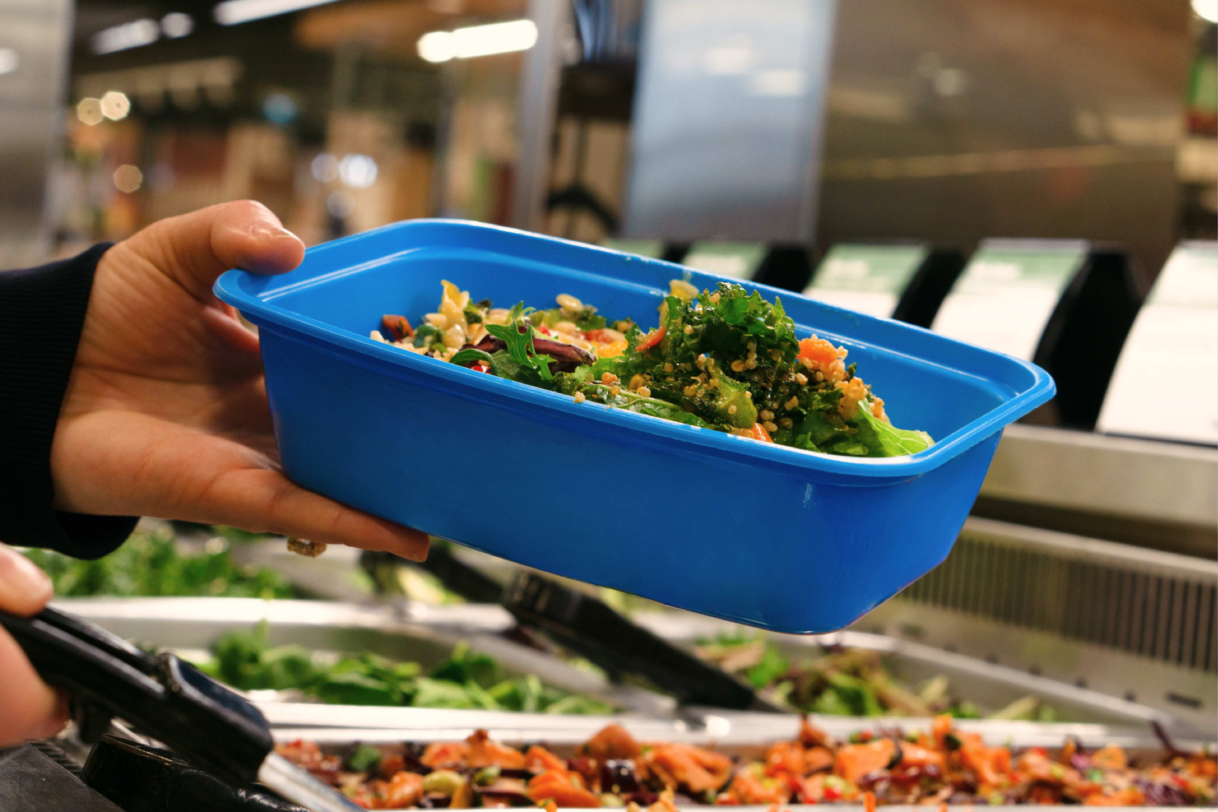A hand holding a blue to-go container at a salad bar with salad inside.