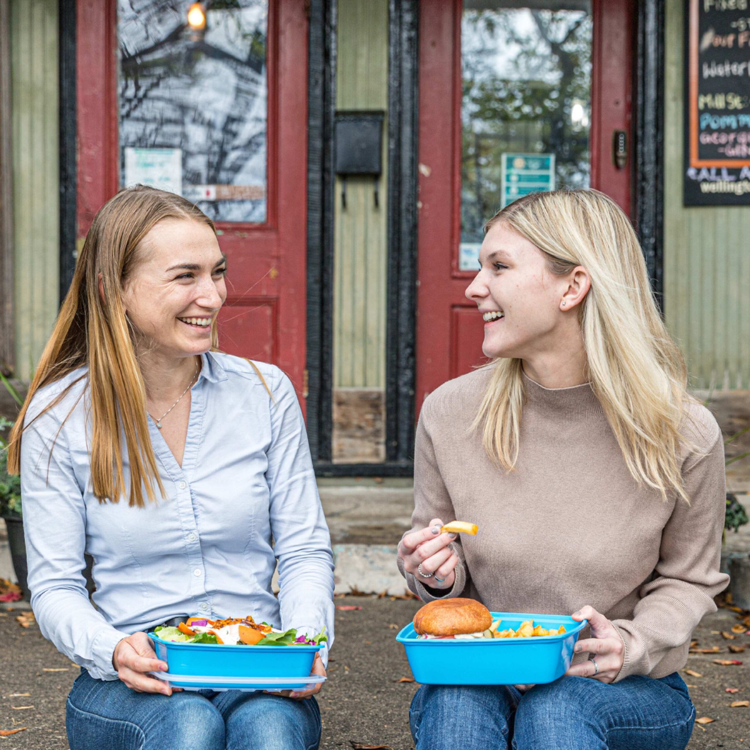 Two women sitting down with blue to-go containers with food on their laps. They are looking at each other and smiling. 