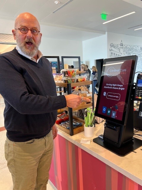 A man with a surprised expression pointing to an ordering screen at a cafe.
